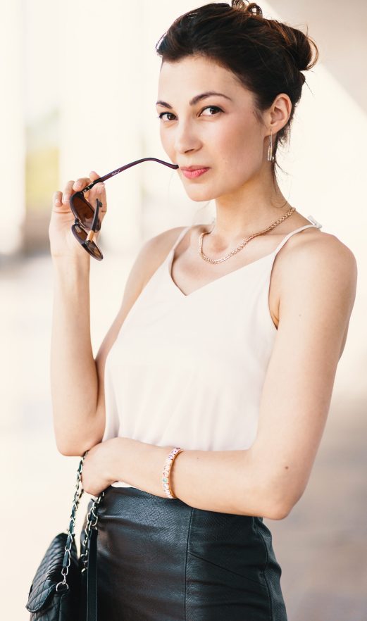 Close-up portrait of young, elegant brunette woman in cream silk Close-up portrait of young, elegant brunette woman in cream silk blouse and leather skirt, touching her lips while holding sunglasses with hand, hair in a bun. Fashion street shot, natural light play